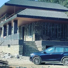 A farmhouse project in Uttarakhand under construction. The design utilizes a raised plinth and deep verandas to respond to the site's topography, while the structure is framed against a backdrop of dense forest.