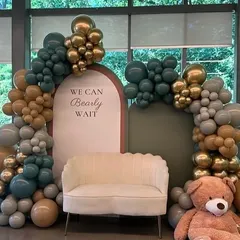 An elegant "We Can Bearly Wait" baby shower decor in a sophisticated palette of sage green, beige, and chrome gold. The setup includes a comfortable scalloped sofa for the mom-to-be, a giant teddy bear, and lush balloon arches for a luxurious feel.