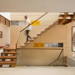 A clean, sculptural staircase with wooden treads and glass railing anchors this space in Kavyam Residence. The double-height wall is accented with patterned tiles, and a laser-cut leaf screen lends a touch of artistry.