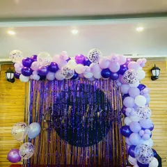 This purple-themed birthday party decor features a large balloon arch with shades of purple, lilac, and confetti balloons. The shimmering purple fringe backdrop creates a fun and glamorous photo booth area.