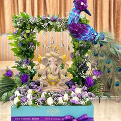 A unique and artistic floral arrangement featuring a Ganesh idol. The idol is placed within a frame of fresh greenery and purple flowers, accompanied by a peacock made of flowers.
