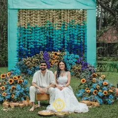 A beautiful setup for a vibrant Haldi and Mehendi ceremony. The backdrop features a stunning gradient of hanging tassels in shades of blue and yellow, complemented by a floral arch of sunflowers and hydrangeas.