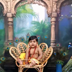 Dressed as little Muruga, this child sits in a peacock-shaped wicker chair, surrounded by a palatial backdrop and real peacock feathers, creating a majestic scene.