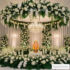 A grand and opulent Ganpati mandap featuring a circular canopy of white hydrangeas, roses, and monstera leaves. The idol is placed centrally under a crystal chandelier, surrounded by pillars of matching white florals for a truly divine look.
