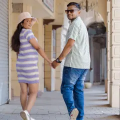 A playful and modern maternity photo of a couple walking through an urban corridor. The mother-to-be, in a casual dress and hat, looks back with a smile while holding her partner's hand, creating a candid and dynamic "follow me" style shot.