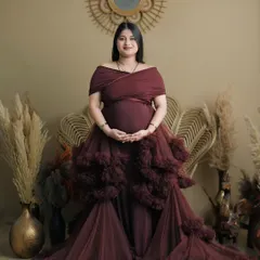 This full-length portrait showcases a stunning ruffled maroon gown. The mother-to-be stands confidently in a bohemian-themed studio setup, creating a look that is both earthy and glamorous.