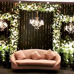 An elegant engagement or reception stage with a dark backdrop that makes the white floral arches and chandeliers pop. The simple pink sofa adds a soft touch of color.