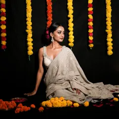 A classic Diwali portrait. The model is seated amidst marigold flower garlands, creating a rich, festive atmosphere that complements her elegant grey saree.