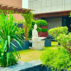 A peaceful corner in a Faridabad garden, where a white Buddha statue sits in meditation. The space is framed by a wooden pergola, a manicured lawn, and carefully sculpted bonsai trees, creating a perfect spot for contemplation.