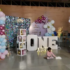 A first birthday party setup with large 'ONE' letters, a sequin wall, and transparent boxes spelling 'BABY'.