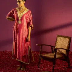 A full-length shot from the Kashidaa campaign, showcasing the complete styled look. The rich pink kaftan is complemented by a traditional choker and a floral hair accessory, set against a dramatic, color-blocked background to create a painterly effect.