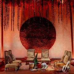 The mandap area for a wedding, featuring a dramatic circular backdrop made entirely of red flowers. The space is defined by hanging floral strings and traditional seating, all bathed in a warm, romantic red light.