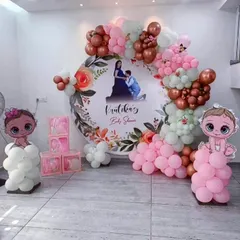 A personalized baby shower decoration featuring a large circular backdrop with the parents-to-be's photo. The setup is framed with a beautiful balloon arch in dusty rose, pink, and white, with cute baby character cutouts.