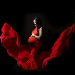A side-profile maternity portrait with a flowing red gown against a dark background. The lighting highlights the baby bump and the beautiful fabric wings.