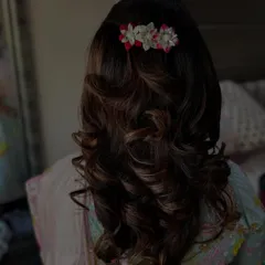 This hairstyle features bouncy curls created with a blow-dry technique on the client's own hair. The half-up twist is adorned with a small floral pin, adding a delicate touch to the voluminous look.