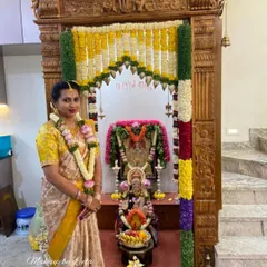 A client celebrating her housewarming ceremony. She is dressed in a traditional silk saree with elegant makeup, standing before her decorated home temple.