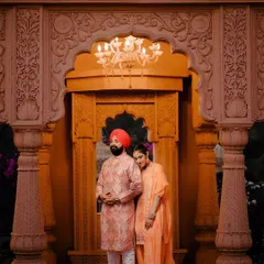 Maha and Ashwen stand within a beautifully carved archway, illuminated by a warm chandelier. The intricate details of the architecture and their traditional outfits create a portrait of pure elegance.