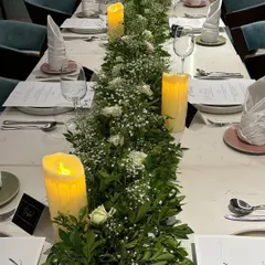 An elegant and fresh table runner for a sophisticated dinner party. We used a lush base of green foliage interspersed with delicate baby's breath, white roses, and tall, flameless candles for a safe and beautiful glow.
