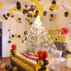 A romantic room decoration with a black and yellow theme. The setup includes "Just Married" foil balloons, a fairy light canopy over the bed, and balloons surrounding the bed frame for a cozy and celebratory feel.
