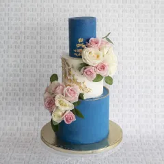 A stunning three-tier wedding cake that combines bold color with classic elegance. The deep blue layers contrast beautifully with the white middle tier, which is accented with gold leaf and a cascade of fresh roses.