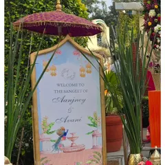 A personalized welcome sign for Avani and Aanjney's Sangeet. The design features beautiful illustrations of peacocks and fountains, topped with a traditional Indian umbrella for a royal touch.
