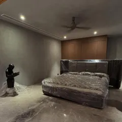 A work-in-progress shot of a bedroom with a moody aesthetic. The space features micro-concrete walls, a custom upholstered bed, and built-in wooden wardrobes, all awaiting final touches.
