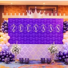 The main stage for Prince Zeeyan's first birthday. A deep purple damask backdrop with his name in ornate lettering is topped with a castle silhouette and framed by beautiful balloon garlands.