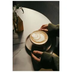 A lifestyle shot focusing on a pair of hands gently holding a cappuccino with beautiful latte art. The soft lighting and moody atmosphere create a cozy and inviting cafe scene.