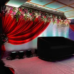 A classic reception stage design featuring a bold red satin drape as the backdrop. I added a thick floral border on top with a mix of pink, white, and purple flowers, creating a rich and elegant look for the couple's seating area.