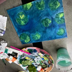 This image shows the work in progress of the koi pond painting. You can see my palette, brushes, and the initial layers of blue and green that form the base of the water scene.