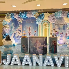 A beautiful Frozen themed stage decoration. This setup includes a custom backdrop with Elsa and Anna, a picket fence, themed plinths, and large marquee letters spelling out the birthday girl's name.