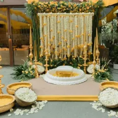 This is an elegant and traditional Haldi ceremony setup. It features a central white seat for the rituals, surrounded by bowls of fresh flower petals, all set against a beautiful backdrop of white and yellow flower strings.