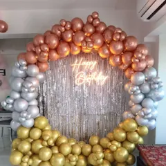 A glamorous circular balloon frame in rose gold, silver, and gold chrome balloons, set against a silver shimmer curtain with a neon sign.