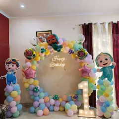 A Cocomelon theme decoration for a first birthday party at home. This setup includes a circular balloon arch in pastel colors, cutouts of the Cocomelon characters, and a light-up number '1'.