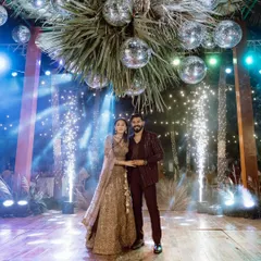 A perfect blend of rustic charm and party glamour for a Sangeet night. The couple poses under a canopy of palm leaves and disco balls, with dramatic sparklers adding to the celebratory mood.