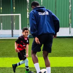 A coach, clearly identified on his hoodie, engages one-on-one with a young player, demonstrating a move and making learning personal and fun.