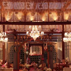 An evening view of a traditionally inspired venue space. The intricate wooden lattice work, grand chandeliers, and rich red carpet create a warm, opulent atmosphere that speaks of heritage and luxury.
