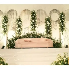 A clean and modern stage design using a white backdrop with minimalist line art. The decor is brought to life with vertical arrangements of white flowers and greenery, and a soft pink sofa adds a subtle touch of color.