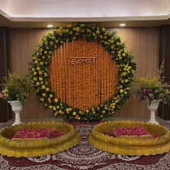 An elegant indoor Haldi setup with two golden lotus-shaped urulis for seating. The backdrop is a large circular arrangement of marigold strings with a "Haldi" sign, set against a simple, modern wooden panel wall.