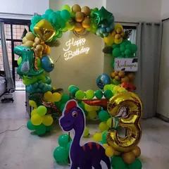 A roaring dinosaur-themed birthday party. The decoration features a green and yellow balloon ring, a neon "Happy Birthday" sign, dinosaur cutouts, and a large number '3' foil balloon.