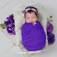 A sleeping newborn is snugly swaddled in a textured purple wrap, lying in a small white basin. The setup is accented with matching purple flowers for a coordinated look.