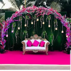 A beautiful reception stage with a cherry blossom theme. The arch of pink and white flowers against a green backdrop, complete with hanging candles, creates a fresh and romantic setting.