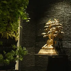 A golden Buddha statue seated under a Bodhi tree is spotlit against a dark, textured wall, creating a dramatic and peaceful corner next to a lush vertical garden.