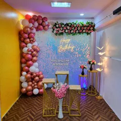 A beautiful party hall decoration featuring a rose gold and white balloon garland against a silver shimmer wall with a floral header.