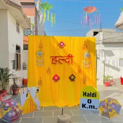 An outdoor Haldi photo corner complete with fun and quirky props. The setup includes a bright yellow backdrop, hanging Rajasthani umbrellas, and a custom "Haldi 0 Km" milestone sign for playful pictures.