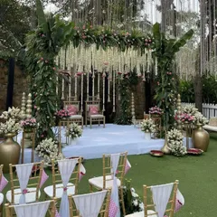 A beautiful and traditional mandap design for an outdoor ceremony. The structure is adorned with lush banana leaves, pink flowers, and hanging tuberose strings, creating a fresh and authentic South Indian wedding vibe.