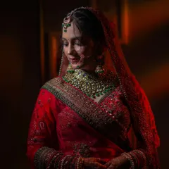 A classic bridal portrait showing the bride in her stunning red lehenga. The soft, dramatic lighting highlights her graceful pose and the richness of her wedding attire.