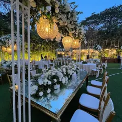 An elegant engagement dinner setup with a white and green theme, featuring long mirrored tables, lush floral runners, and beautiful overhead chandeliers.