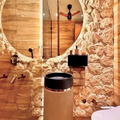 This view shows how the different textures in the powder room work together. The reflection in the large circular mirror captures the ceiling fan and adds a sense of depth, while the lighting accentuates the ruggedness of the stone wall.