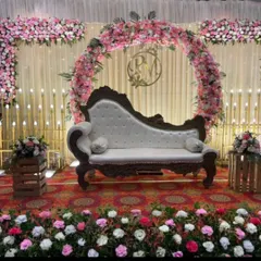 This stage setup features a circular floral arch with pink and white roses and a custom monogram of the couple's initials. The vintage-style chaise lounge adds a touch of royal elegance.